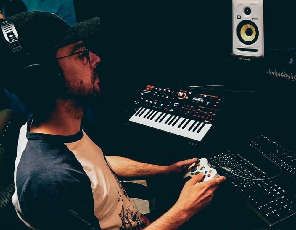 David at his desk with headphones on, a game controller in his hands and a synth in front of him.